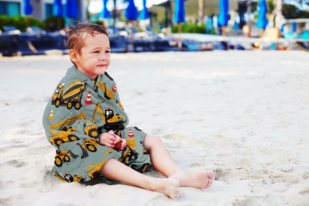 A happy child wrapped in a soft organic cotton towel featuring an exciting construction truck design with yellow excavators, dump trucks, and traffic cones. The towel is made from high-quality, eco-friendly organic cotton, offering both comfort and absorbency for kids after a fun day at the beach or pool. The playful construction-themed pattern adds a touch of adventure, making it a perfect choice for little ones who love trucks. Durable, lightweight, and gentle on the skin, this towel is an ideal addition 