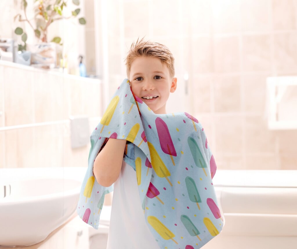 A happy child using a soft organic cotton towel featuring a playful design of colorful ice cream popsicles in pink, yellow, and mint green on a light blue background. The towel is soft, absorbent, and perfect for kids, ensuring comfort after a bath or shower. The cheerful popsicle pattern adds a fun element, making it an exciting option for young ones. Crafted from eco-friendly organic cotton, it provides durability and a gentle touch, making it both a practical and delightful addition to your child’s bath 